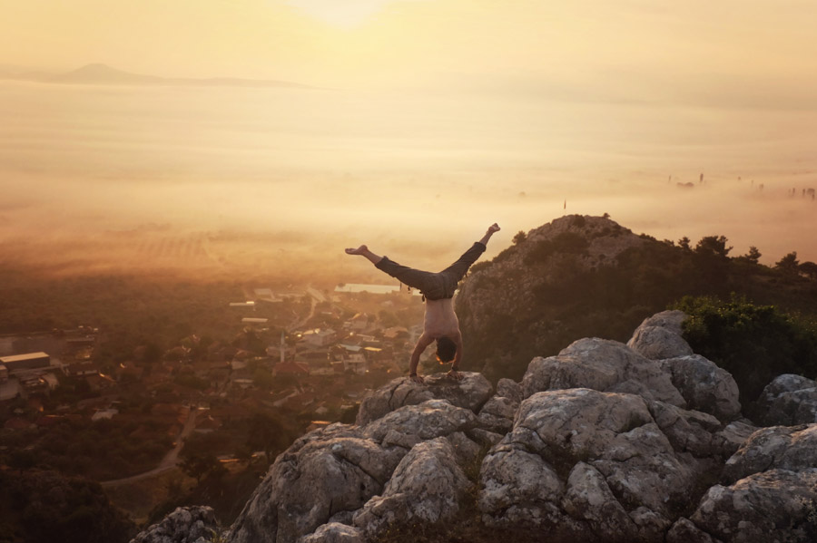 フリー写真 山の上の岩で逆立ちする男性と朝陽の風景